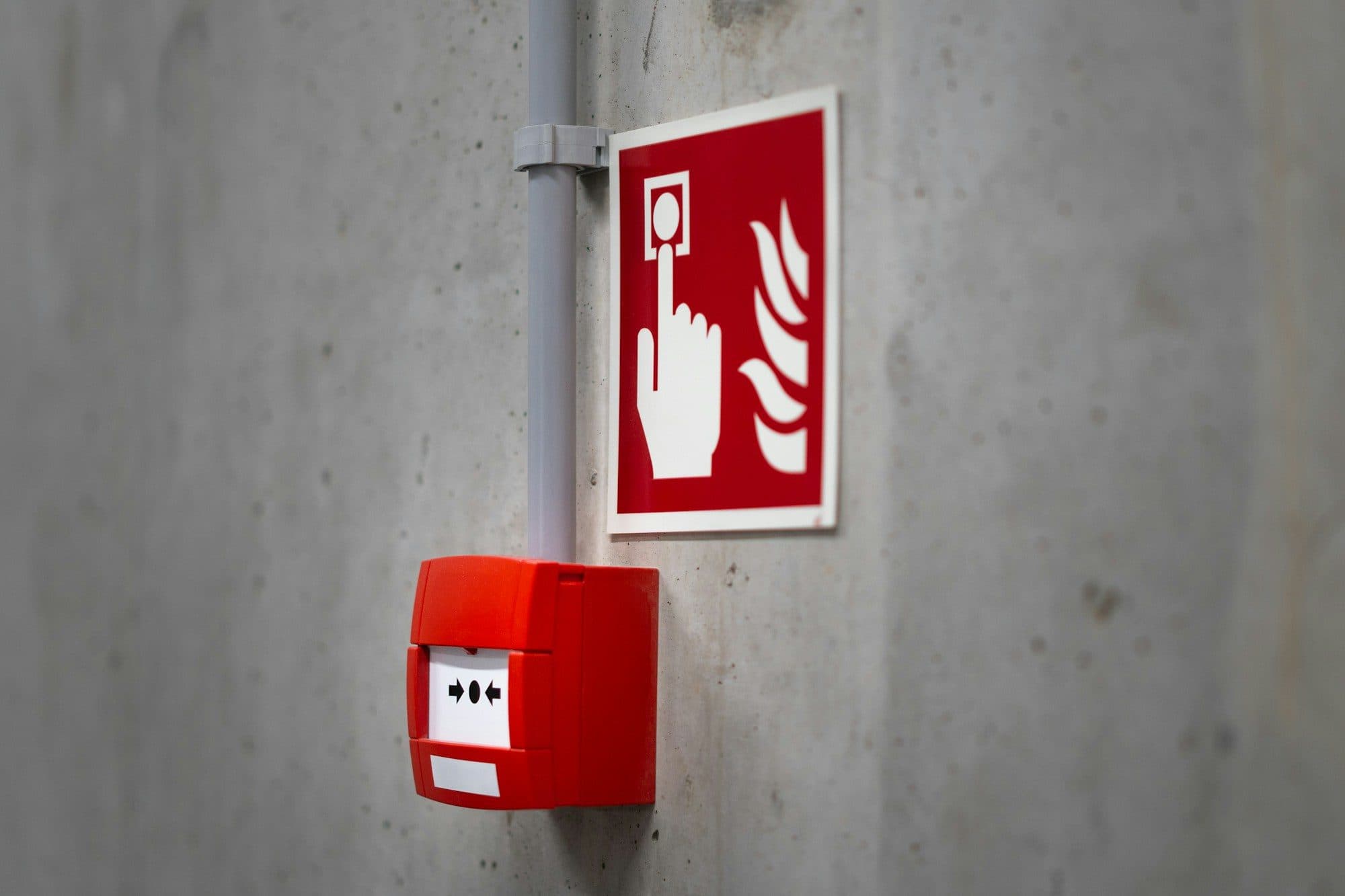 Red fire alarm call point mounted on a concrete wall below a fire alarm sign